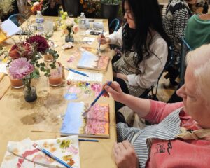 Several people sit at a table painting floral landscapes on canvases, surrounded by paint supplies and vases of flowers.