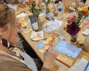 A woman paints a landscape on canvas at a table covered with art supplies, paint palettes, water bottles, and flower arrangements.