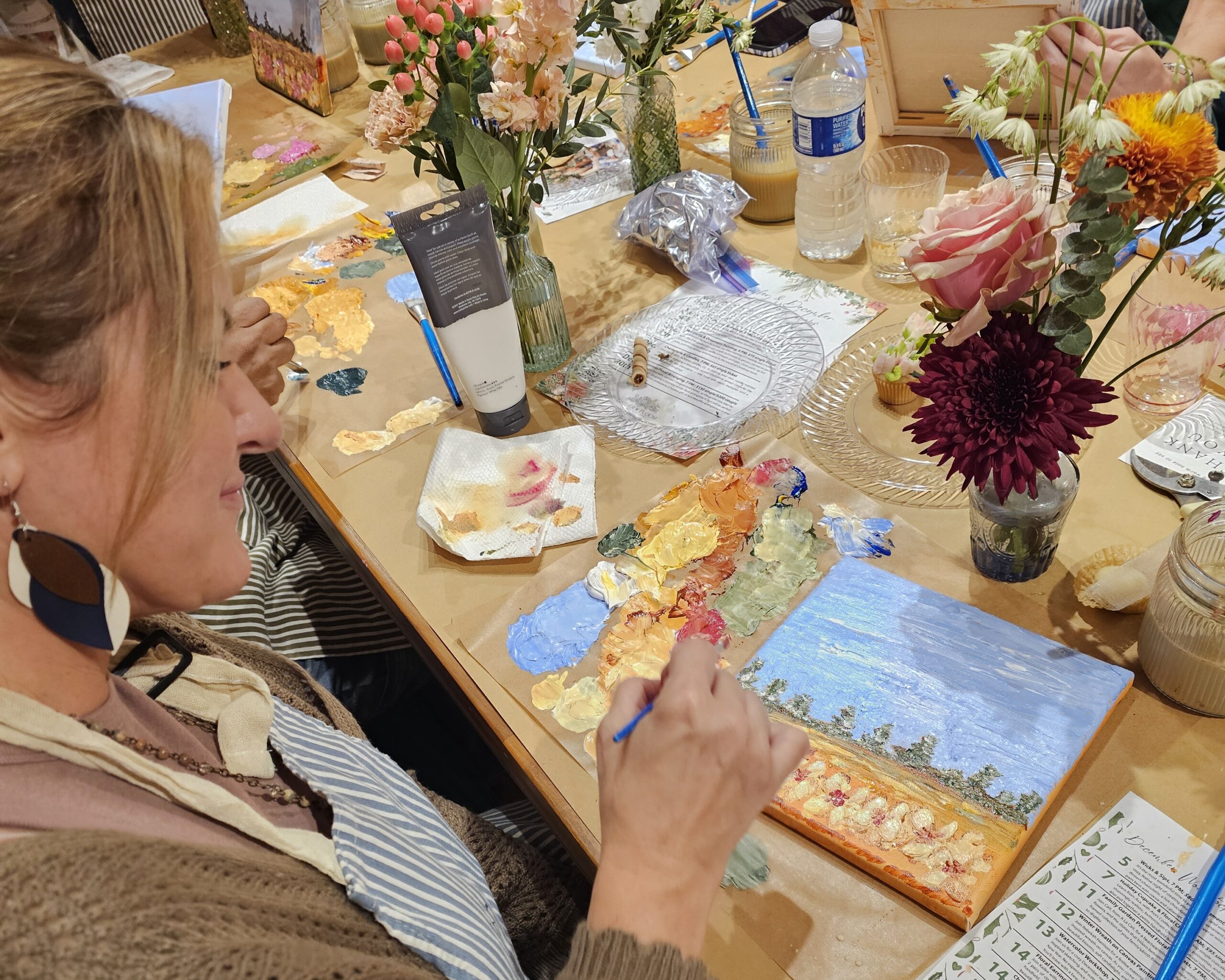 A woman paints a landscape on canvas at a table covered with art supplies, paint palettes, water bottles, and flower arrangements.