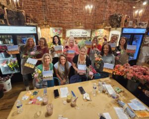 A group of women pose indoors holding floral arrangements and paintings at a flower shop workshop called The Bloom Bar.