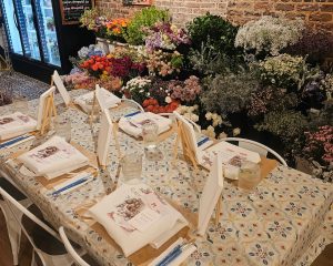 A table set with easels, paper, and art supplies stands in front of shelves filled with various fresh flowers in a well-lit room.