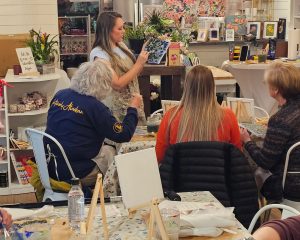 A woman stands at the front of a room demonstrating a painting technique to a seated group, who observe with blank canvases and supplies on tables.