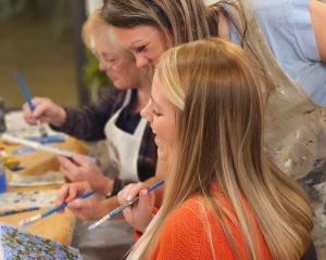 Three women sit at a table painting on canvases, each holding a paintbrush and wearing aprons, focused and engaged in their artwork.