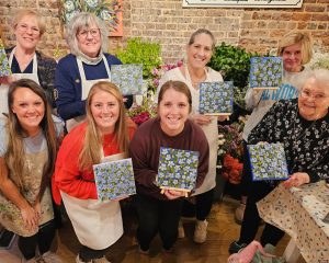 A group of seven women pose indoors, each holding a square painting of blue flowers, with floral arrangements and a brick wall in the background.