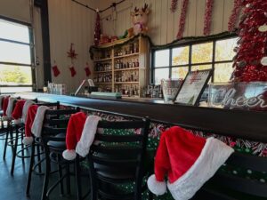 A bar decorated for Christmas with Santa hats on chairs, red tinsel, a reindeer plush on a shelf, and festive signs, with sunlight coming through large windows.