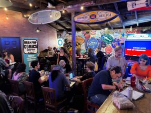 A busy bar or café with people eating, drinking, and working on laptops. The walls are decorated with vintage signs and posters.
