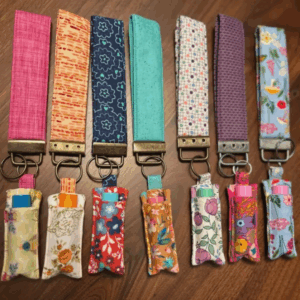 Five fabric keychains with matching chapstick holders, each in different colorful patterns, displayed on a wooden surface. Each holder contains a tube of lip balm.
