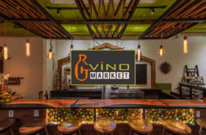 A modern wine bar with a wooden counter, stools, decorative lighting, wine bottles, and a large "Vino Market" sign displayed at the center.