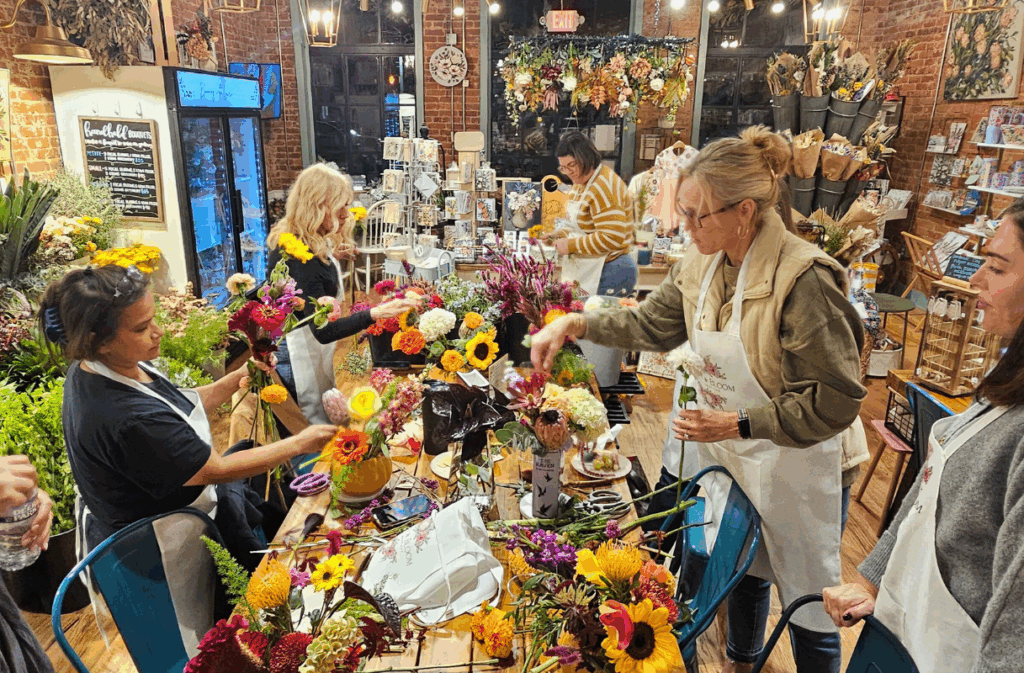 People arranging colorful flowers on a table in a flower shop, wearing aprons, surrounded by various floral displays and shop decor.