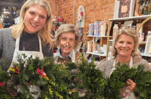 Three women indoors, each holding a decorated holiday wreath with greenery, pinecones, and ribbons, standing in front of shelves with cards and gift items.