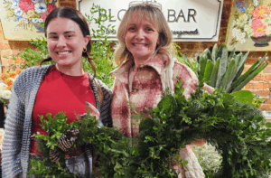 Two women stand indoors holding greenery wreaths, with a backdrop of plants and a sign reading "BLOOM BAR" partly visible behind them.