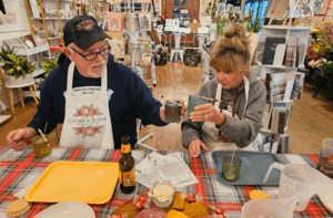 An older man and woman wearing aprons sit at a table with snacks, drinks, and craft supplies, clinking glasses together in a brightly lit craft store.
