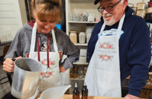 Two people wearing aprons pour liquid from a metal jug into a container on a table, with bottles, jars, and shelves visible in the background.