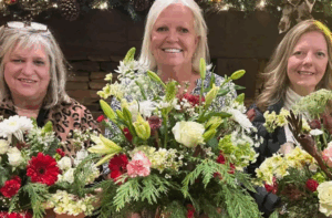 Three women standing indoors hold large floral arrangements with a mix of red, white, and green flowers and greenery. A festive garland decorates the background.