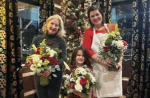 Three people, two adults and one child, stand indoors in front of a decorated Christmas tree, each holding a bouquet of flowers and smiling at the camera.