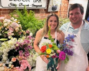 Two people wearing aprons stand in front of a display of assorted flowers, holding a colorful bouquet; a “Bloom Bar” sign is visible behind them.