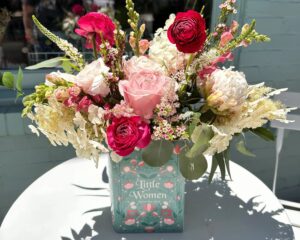 A floral arrangement with pink, red, and white flowers is displayed in a vase designed to look like the book "Little Women" on a white table.