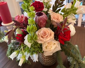 A floral arrangement in a textured glass vase with roses, protea, carnations, snapdragons, and greenery on a wooden table. A red candle is in the background.