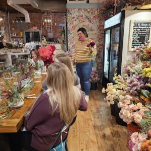 A group of women sit at a wooden table arranging flowers while another woman stands holding a bouquet in a flower shop decorated with floral displays.