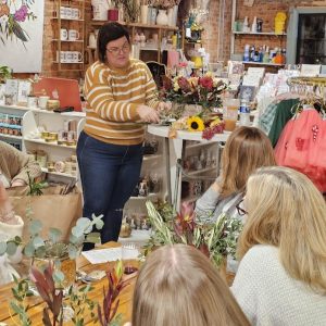 A woman in a striped sweater demonstrates flower arranging to a seated group in a shop filled with gifts and floral supplies.