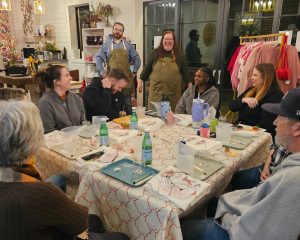A group of people sit around a table with drinks and food, engaged in conversation and laughter, with two people standing and smiling in the background.