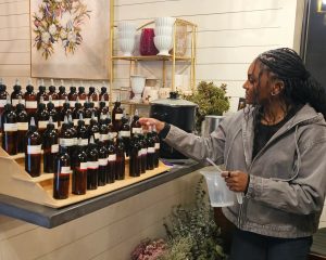 A person selects a brown glass bottle from a display of essential oils in a store, holding a clear measuring pitcher.