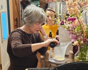 An older woman pours liquid from a brown bottle into a measuring cup on a scale, while a younger woman watches. Flowers and brown bottles are on the wooden counter.
