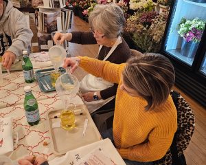 Two women seated at a table pour liquid into glass containers during a workshop inside a flower shop. Bottles of sparkling water and flowers are visible on the table and in the background.