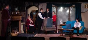 Six people are on stage in a living room set—three standing, three seated—surrounded by formal decor. This scene could be part of a play inspired by “26 Acworth-y Things to do in 2026,” capturing the city's vibrant community spirit.