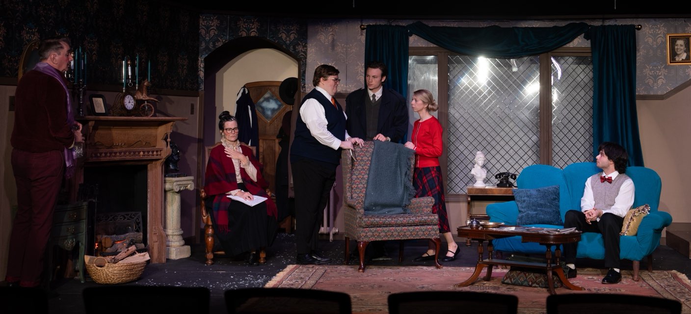 Six people are on stage in a living room set—three standing, three seated—surrounded by formal decor. This scene could be part of a play inspired by “26 Acworth-y Things to do in 2026,” capturing the city's vibrant community spirit.