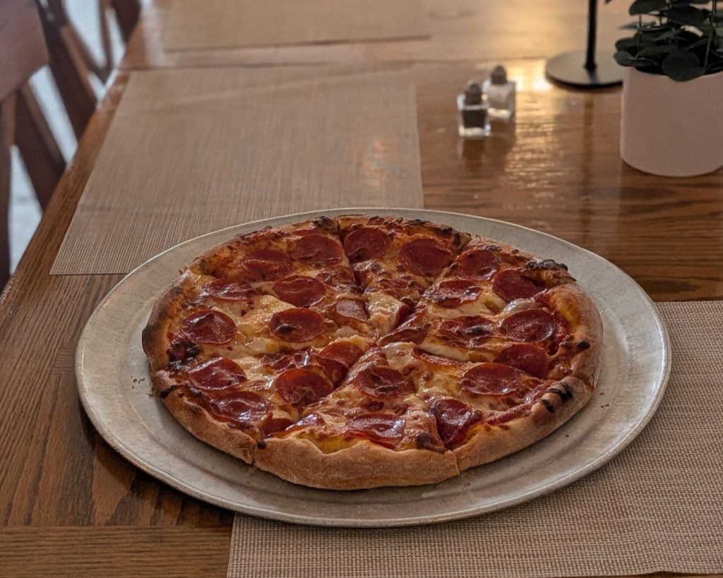 A pepperoni pizza on a round metal tray sits on a wooden table set with placemats, a small plant, and salt and pepper shakers.