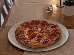A pepperoni pizza on a round metal tray sits on a wooden table set with placemats, a small plant, and salt and pepper shakers.