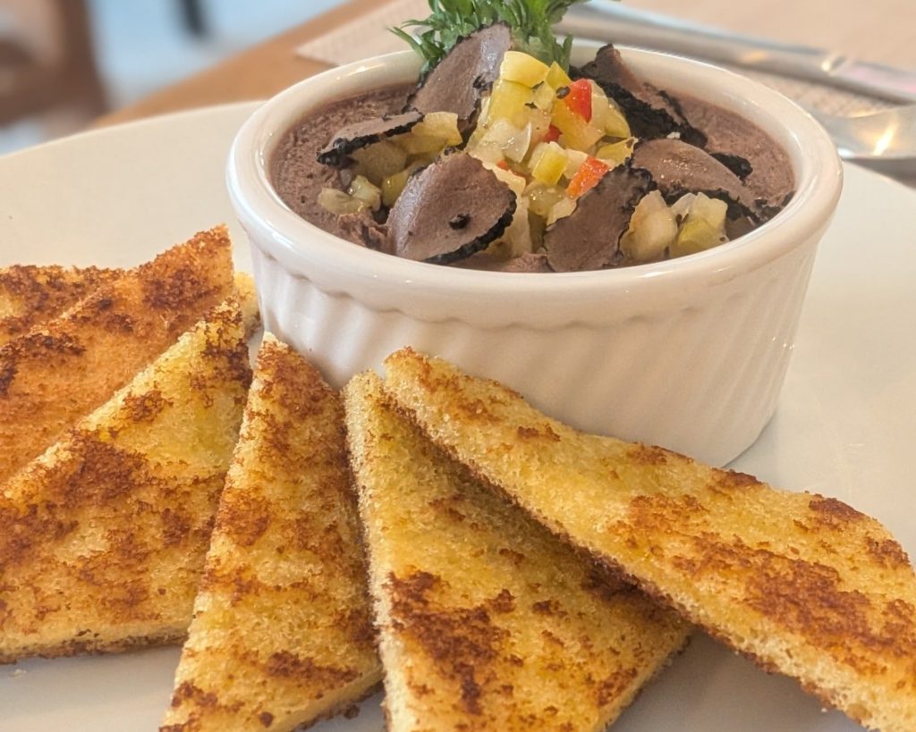 A white ramekin filled with pâté, garnished with black truffle slices and diced vegetables, is served with several toasted bread triangles on a white plate.