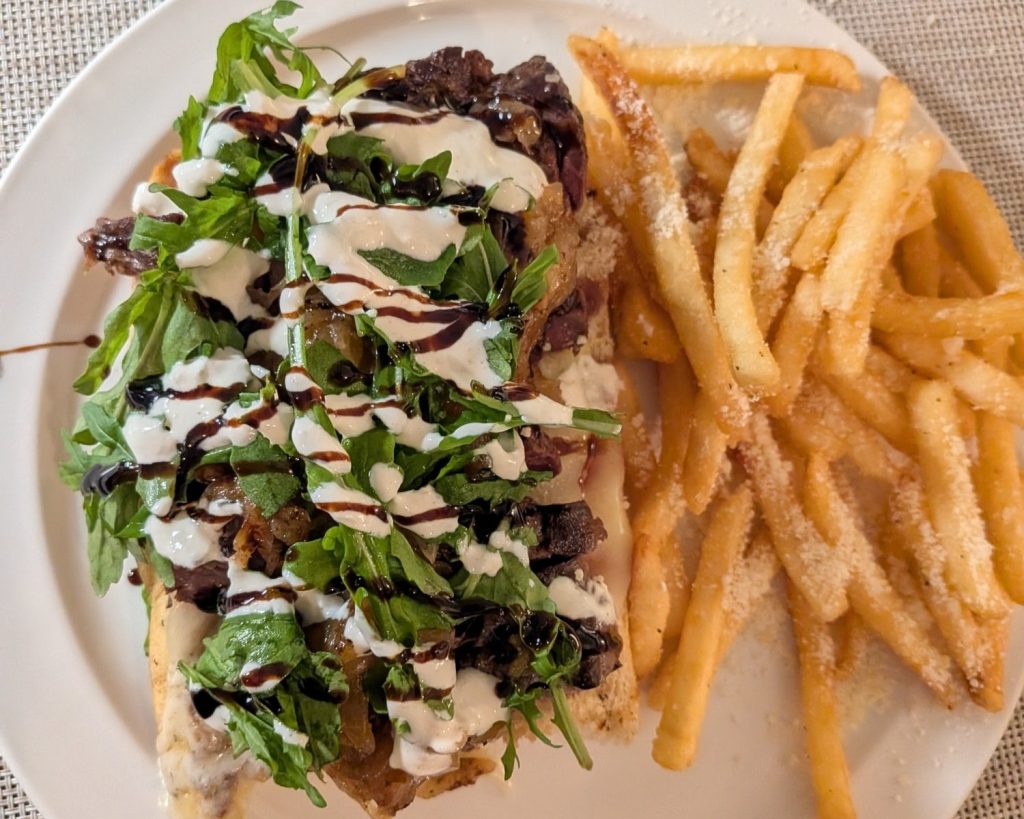 A plate with a sandwich topped with greens, drizzled sauces, and a side of seasoned French fries.