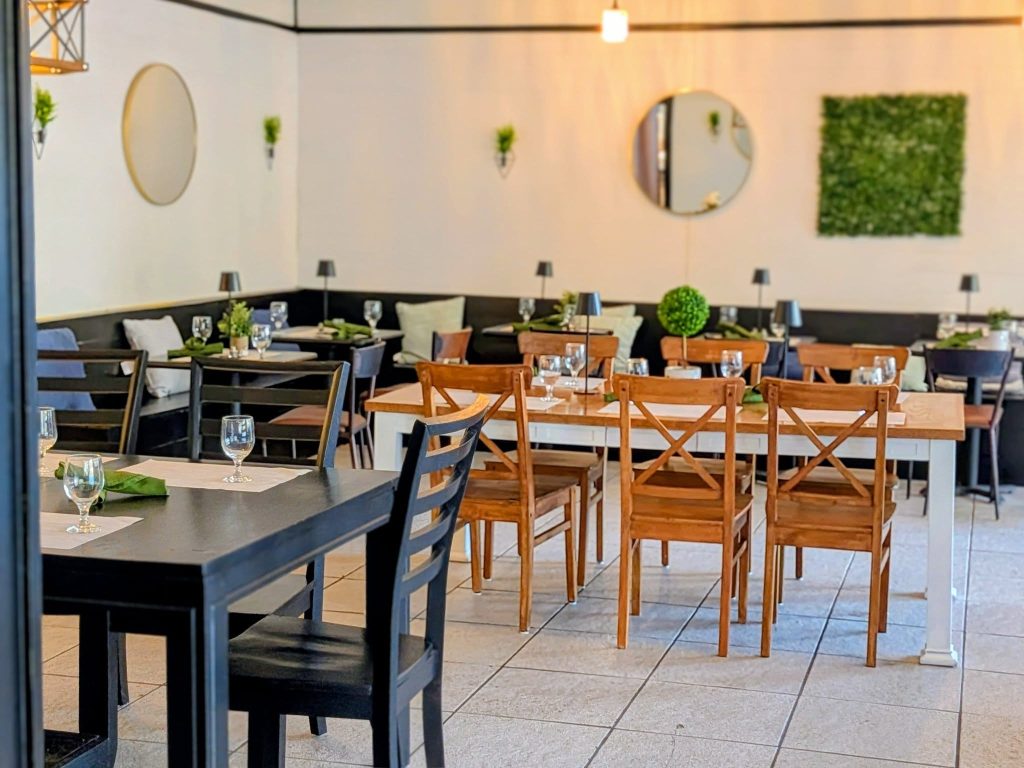 A modern restaurant dining area with wooden tables and chairs, set with glassware and napkins, featuring minimalist wall decor and potted plants.