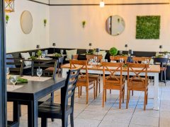 A modern restaurant dining area with wooden tables and chairs, set with glassware and napkins, featuring minimalist wall decor and potted plants.