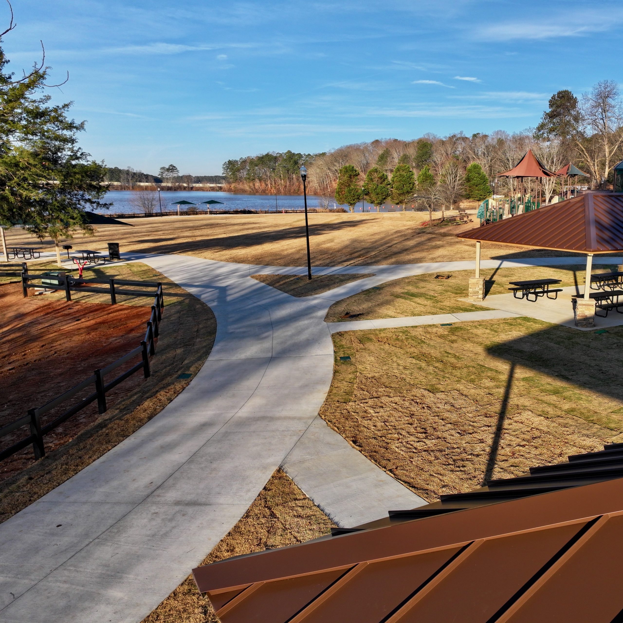 A paved walkway curves through a park with picnic tables, a playground, and a covered pavilion near a lake with trees in the background—perfect for exploring some of the 26 Acworth-y Things to do in 2026.