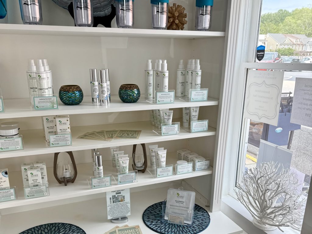 Shelves display various skincare products in white bottles and boxes, alongside decorative items, near a sunny window overlooking a parking lot and street.