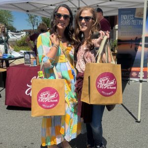 Two women stand outdoors under a canopy, each holding a large brown tote bag with a pink "Welcome to Atascadero" sticker—perhaps collecting ideas for the "26 Acworth-y Things to do in 2026.