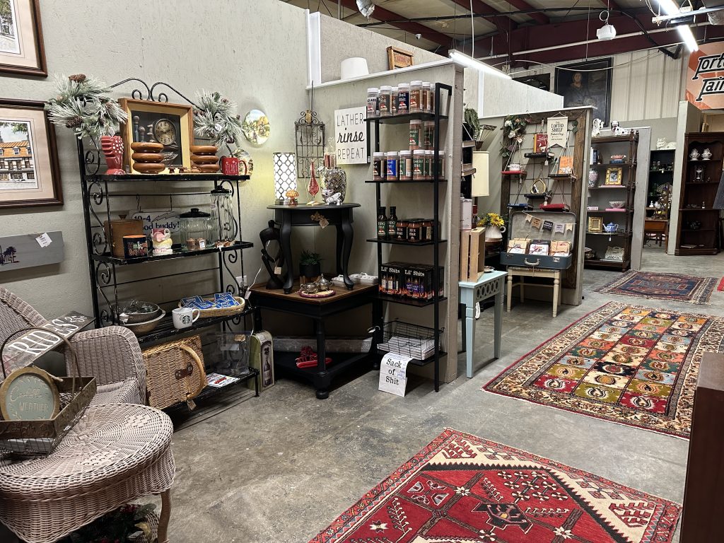 A shop aisle with shelves displaying assorted home decor, spices, gifts, rugs, and wicker chairs on a polished concrete floor.
