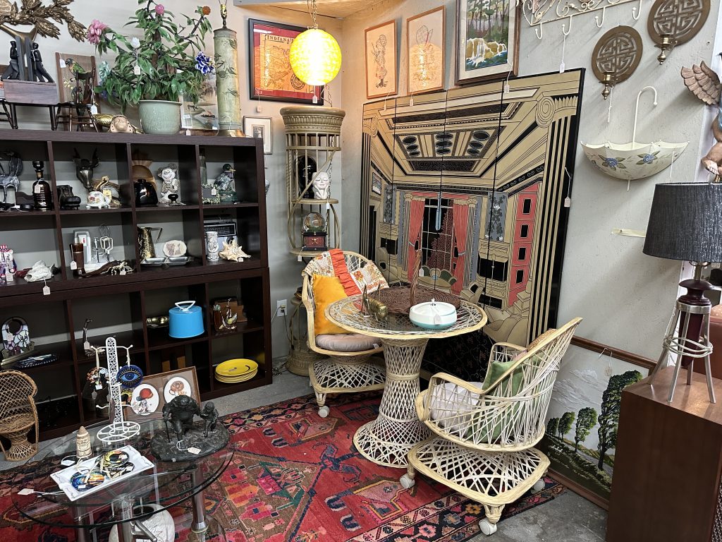 Eclectic vintage shop corner with wicker furniture, assorted decorative items on shelves, framed art, potted plants, and a rug with a red pattern.