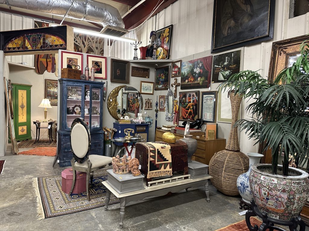 An antique store interior displays vintage furniture, paintings, rugs, and decorative objects, including trunks, lamps, mirrors, plants, and pottery, arranged in a cluttered yet organized setting.