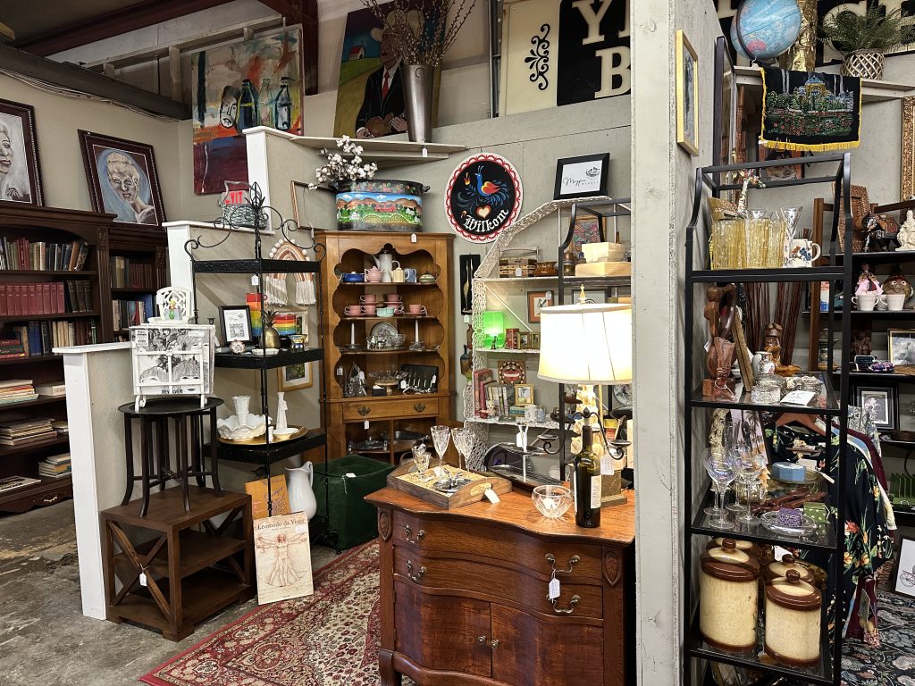 A vintage store displays shelves and cabinets filled with books, glassware, ceramics, lamps, art, and decorative objects in a cluttered, eclectic arrangement.