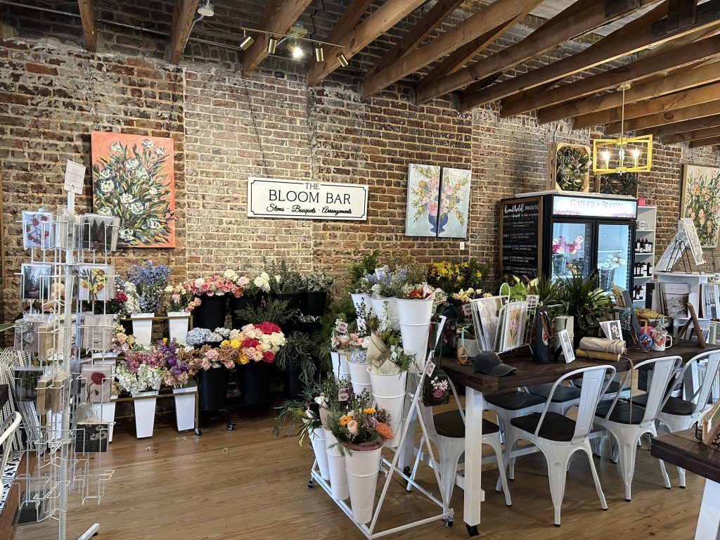 A flower shop interior with brick walls, floral arrangements, artwork, a display rack, a refrigerator, and tables with chairs.