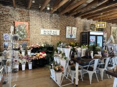A flower shop interior with brick walls, floral arrangements, artwork, a display rack, a refrigerator, and tables with chairs.