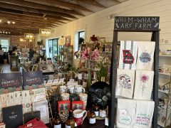 A gift shop interior displays assorted cards, towels, mugs, books, candles, and art under a sign reading "Whim-Wham Art Farm.