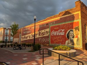 A brick wall features vintage advertisements for Mother's Self-Rising Flour and Coca-Cola, with a bench and landscaped plaza in the foreground—one of the 26 Acworth-y Things to do in 2026.