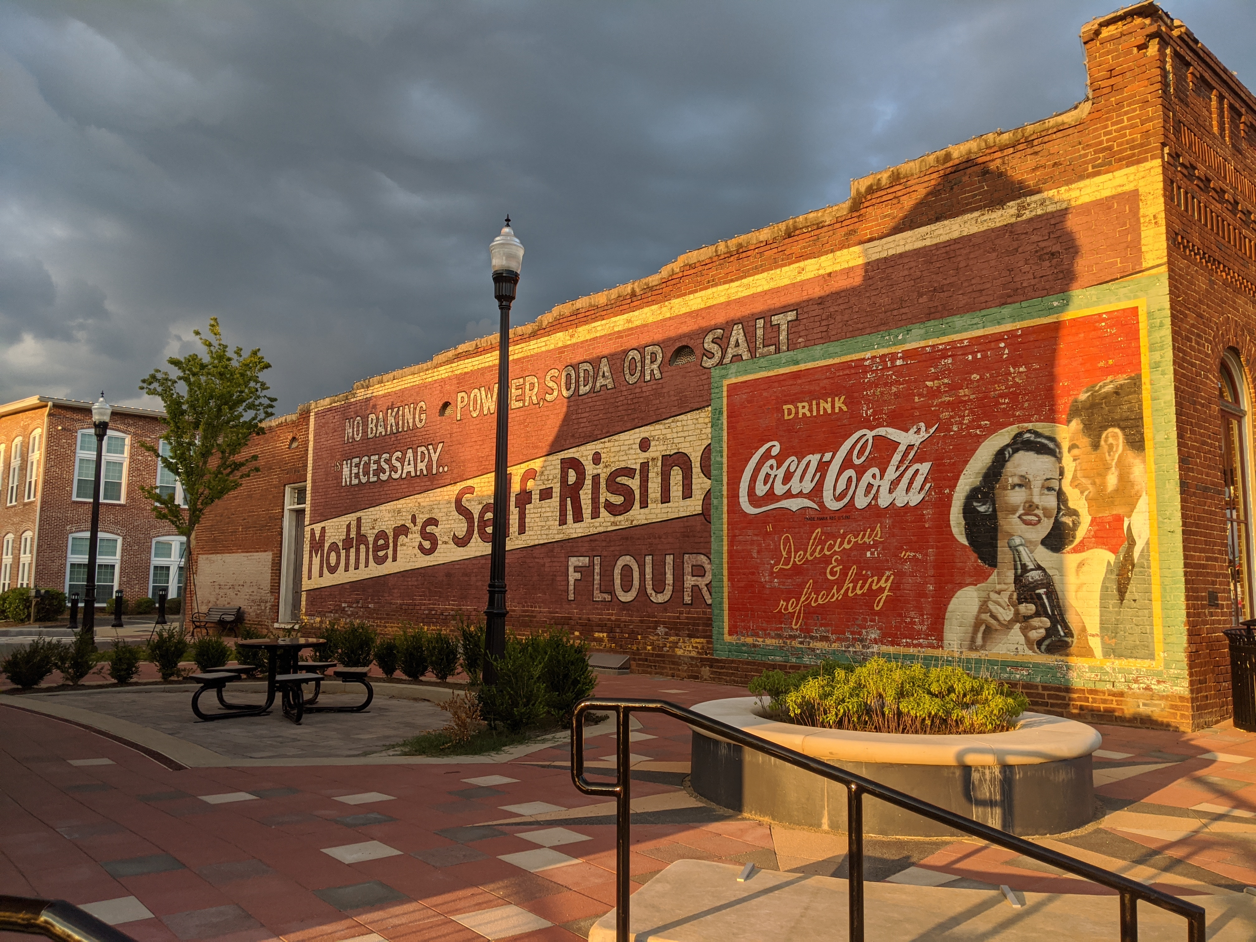 A brick wall features vintage advertisements for Mother's Self-Rising Flour and Coca-Cola, with a bench and landscaped plaza in the foreground—one of the 26 Acworth-y Things to do in 2026.