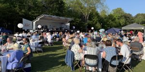 People sit at round tables on a lawn during an outdoor event, listening to a band perform on stage; trees and tents are in the background—one of the 26 Acworth-y Things to do in 2026.