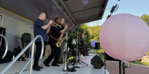 A band performs on an outdoor stage with a saxophonist, trumpeter, and singer; a large pink balloon is visible in the foreground—just one highlight from the 26 Acworth-y Things to do in 2026.